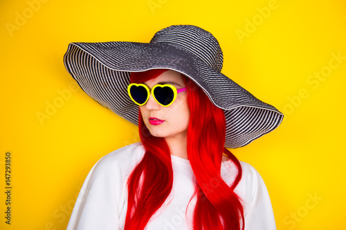 Attractive red-haired young woman in sunglasses and hat on yellow background