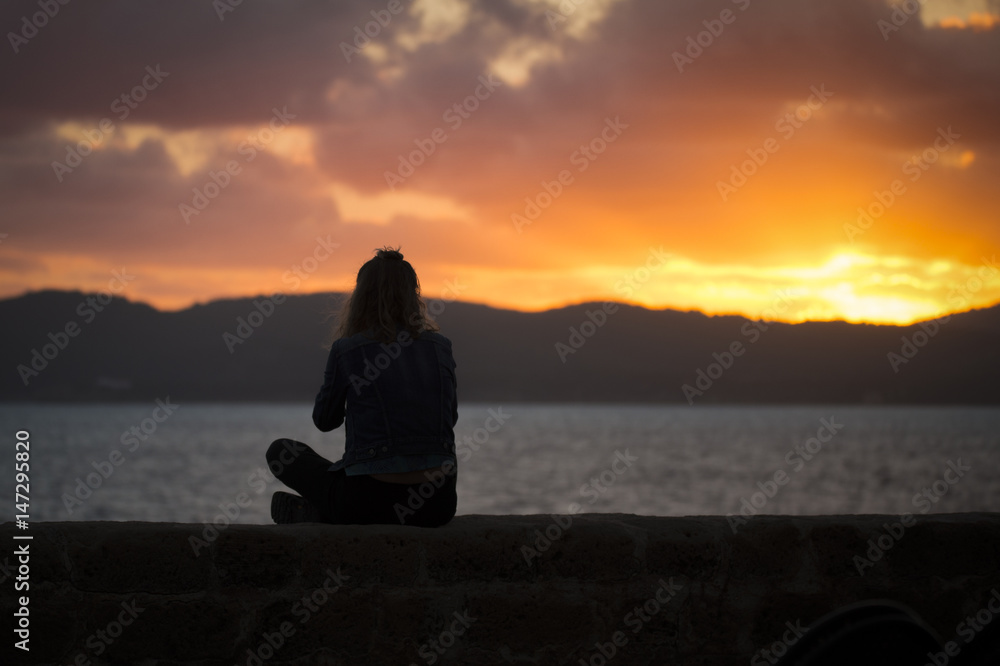 Fototapeta premium A silhouette of a girl is relaxing at sunset enjoying the view of the sea from the ancient walls of a fortification in Alghero at sunset,