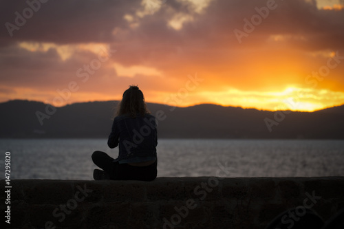 A silhouette of a girl is relaxing at sunset enjoying the view of the sea from the ancient walls of a fortification in Alghero at sunset,