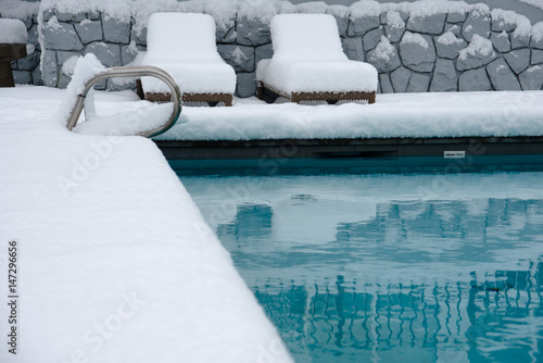 An outdoor swimming pool. the area around it and the lounging chairs  covered in over half a foot, 20cm, of snow.