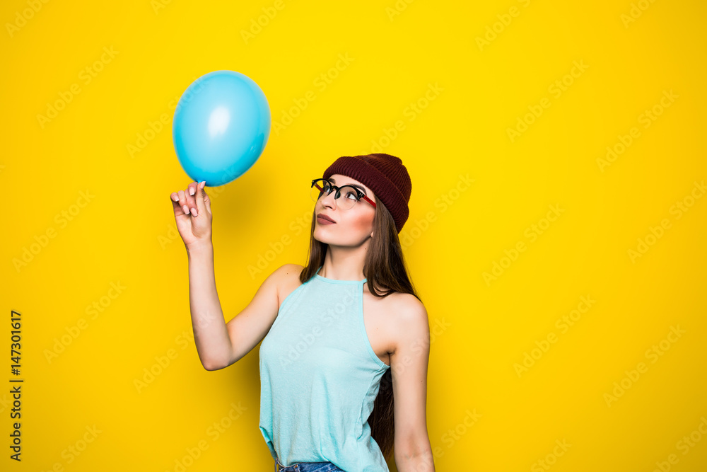 Happy hipster girl in red hat and jeans with blue balloon on yellow wall Happy hipster girl in red hat and jeans with blue balloon on yellow wall