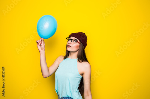 Happy hipster girl in red hat and jeans with blue balloon on yellow wall
