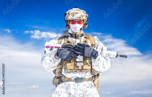 Army serviceman in winter camo somewhere in the Arctic. He wears chest rig and huge backpack  suffers from the extreme cold and strong wind  but endures while mission continues. Blue sky background