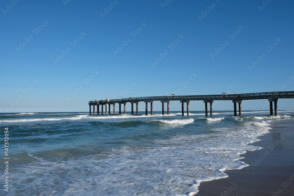 St Johns County Ocean Pier stretching to the horizon, sea lapping on the sandy shore of the beach in St Augustine, Florida, USA on a sunny afternoon. Clear blue sky with no clouds, St Augustine Beach.