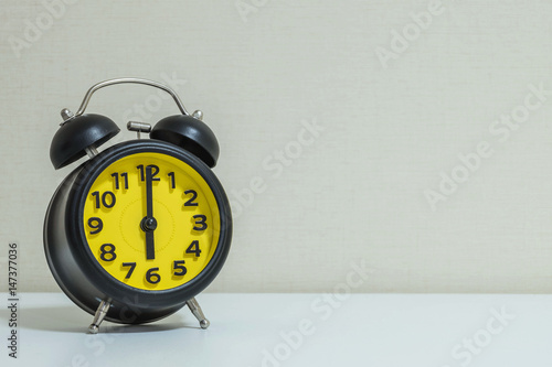 Closeup yellow and black alarm clock for decorate in 6 o'clock on white wood desk and cream wallpaper textured background with copy space