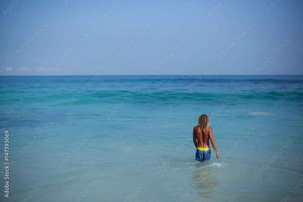 Horizontal panoramic portrait of an attractive curly wet man in blue shorts and with fit naked torso is going into the ocean and, located on the right side of the frame. View from back