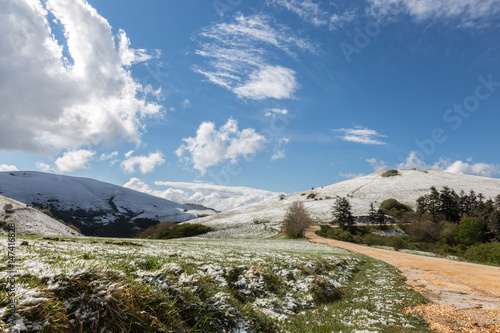 A mountain scenery with green grass and snow, under a blue sky with white clouds of various shapes