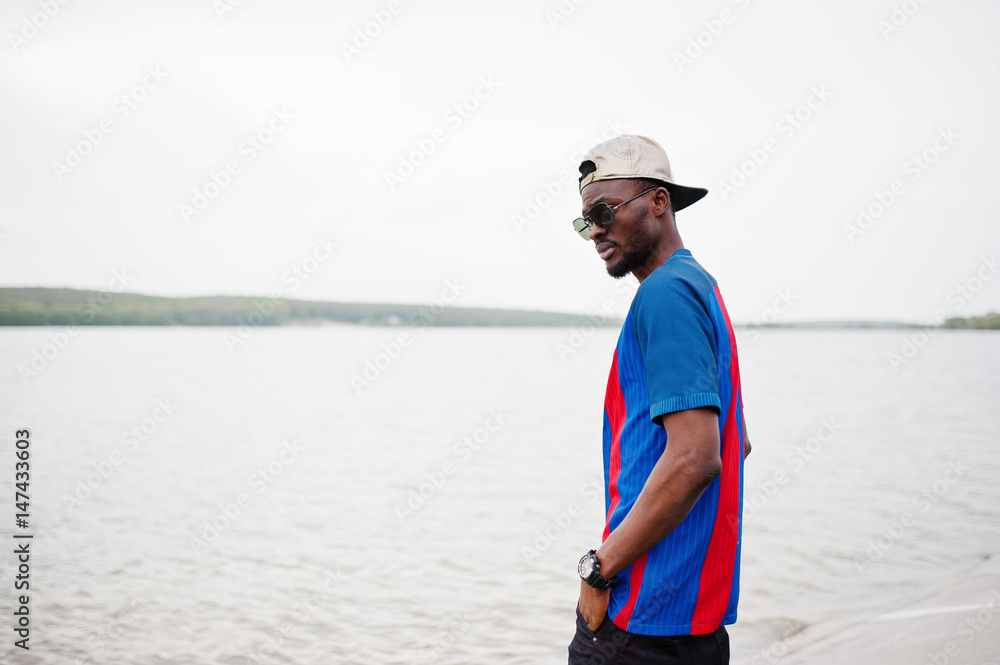 Stylish african american boy wear at cap, football t-shirt and sunglasses stay against lake beach. Black sports man portrait. Stylish african american boy wear at cap, football t-shirt and sunglasses stay against lake beach. Black sports man portrait.