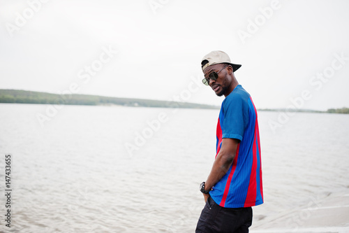 Stylish african american boy wear at cap, football t-shirt and sunglasses stay against lake beach. Black sports man portrait. © AS Photo Family
