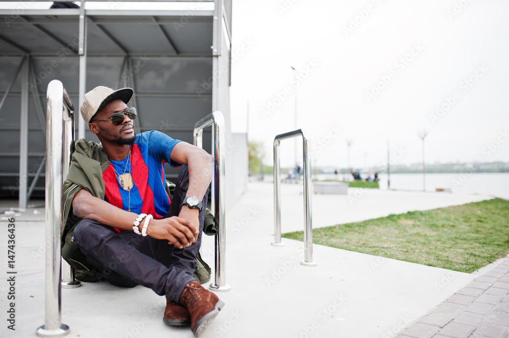 Fototapeta premium Stylish african american boy wear at cap, football t-shirt and sunglasses sitting near steel railings. Black sports man portrait.