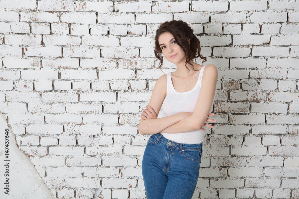 Beautiful  Woman is looking at camera  while standing with crossed arms against white brick wall.