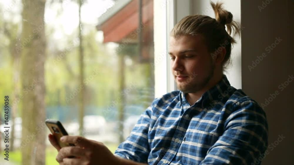 Happy man with mud bun sitting next to the window and texting on smartphone
Happy man with mud bun sitting next to the window and texting on smartphone