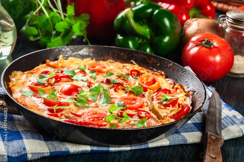 Frittata made of eggs, sausage chorizo, red pepper, green pepper, tomatoes, cheese and chili in a pan on wooden table