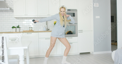 Beautiful young woman in sweat short dancing expressively while listening to music with headphones in kitchen alone. 