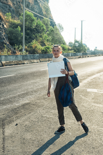 Young woman hitchhiker standing on the road. Tired happy girl with blue backpack holding a white blank cardboard sign. Hitchhiking at sunset or sunrise