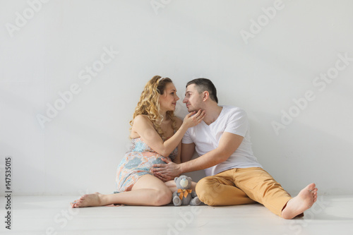 Pregnant family sitting on the floor embracing, holding hands, white room