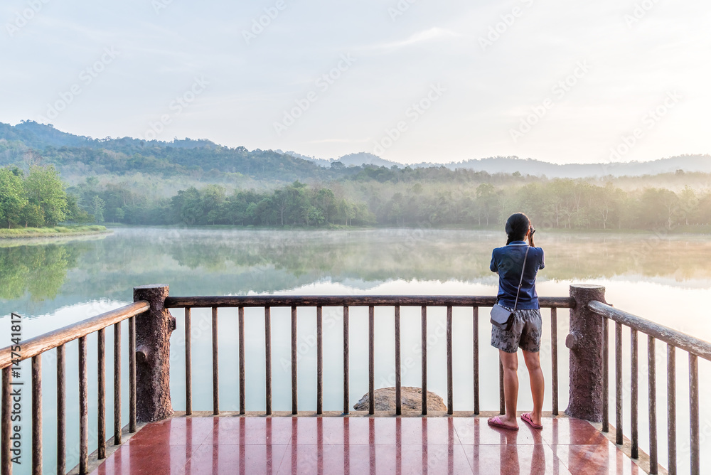 Back view of young woman taking photograph on the wooden balcony and looking at lake on morning.