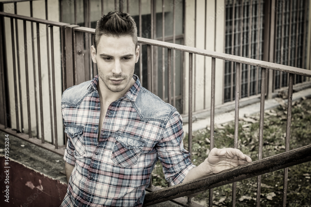 An attractive young man sitting on ground outdoor in public park against metal fence, wearing jeans and shirt An attractive young man sitting on ground outdoor in public park against metal fence, wearing jeans and shirt