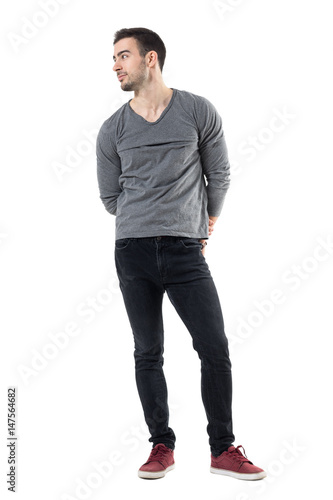 Happy young smiling casual man with hands behind back looking away. Full body length portrait isolated over white studio background.