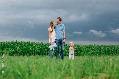 Family standing in front of the cornfield and thunder sky. Father hugs mother with teddybear and daughter. Green grass