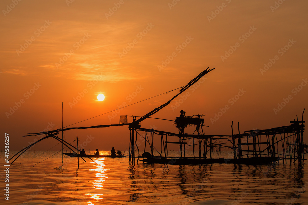 Asian fisherman on wooden boat casting a net for catching freshwater fish in nature river in the early morning with sunrise