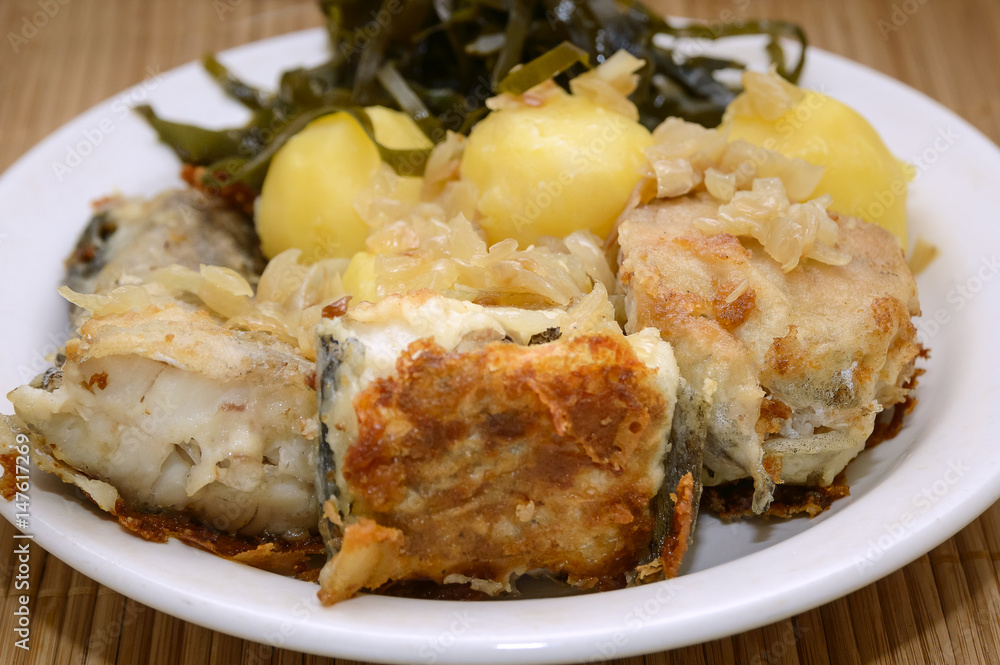 Fried fish, boiled potatoes and sea kale lies on a plate on a bamboo napkin. Close-up, selective focus. Fried fish, boiled potatoes and sea kale lies on a plate on a bamboo napkin. Close-up, selective focus.