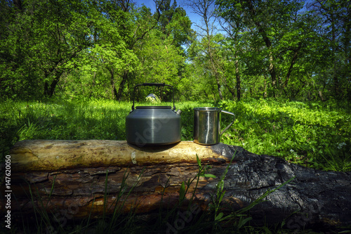 Kettle and mug on the charred log in sunny spring day with forest and tent background