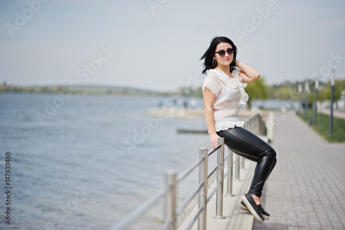 Portrait of  brunette girl on women's leather pants and white blouse, sunglasses, against iron railings at beach. © AS Photo Family