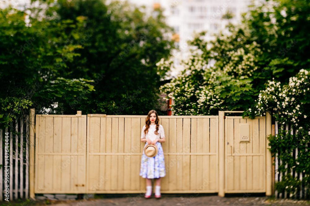 Naklejka premium Old fashioned russian stylish girl. Cutie young in vintage retro long skirt, white top and curly red hair and straw hat posing for camera with fence and green trees on background. Pin up summer style