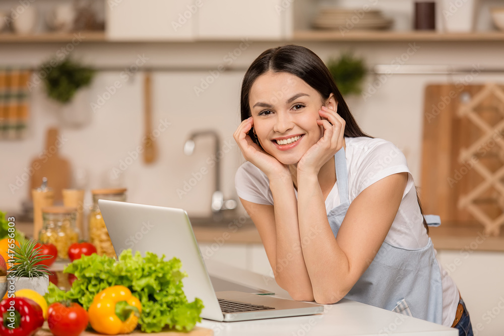 Young Woman in kitchen with laptop, looking recipes, smiling. Food blogger concept