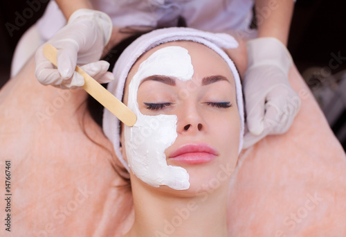 The doctor is a cosmetologist for the procedure of cleansing and moisturizing the skin, applying a mask with stick to the face of a young woman in beauty salon.Cosmetology and professional skin care.