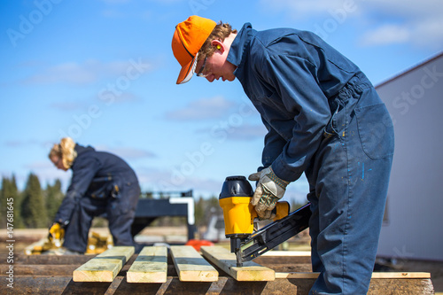 A teenage boy and a blurred teenage girl building outdoors with a power tool wearing protective industrial clothing