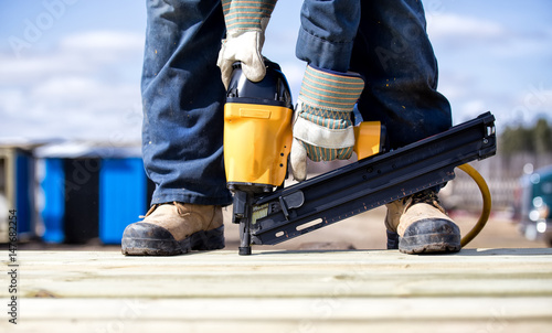 Close up of legs and feet in steel toe boots and arms of man in overalls and work gloves using an air nailer on wood boards outdoors in summer