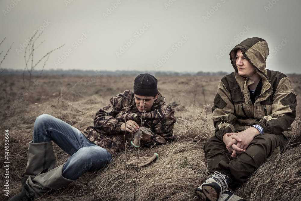 Young men with serene and peaceful expression in camouflage having rest in rural field relaxing on dry grass after long hunting day with dramatic sky Young men with serene and peaceful expression in camouflage having rest in rural field relaxing on dry grass after long hunting day with dramatic sky