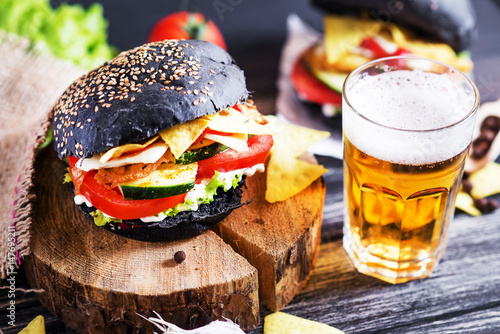 Black burger with chicken, lettuce, mayonnaise, ketchup, tomato, cucumber, cheese, corn chips and a light beer on a dark wooden background, a roll with cuttlefish ink 