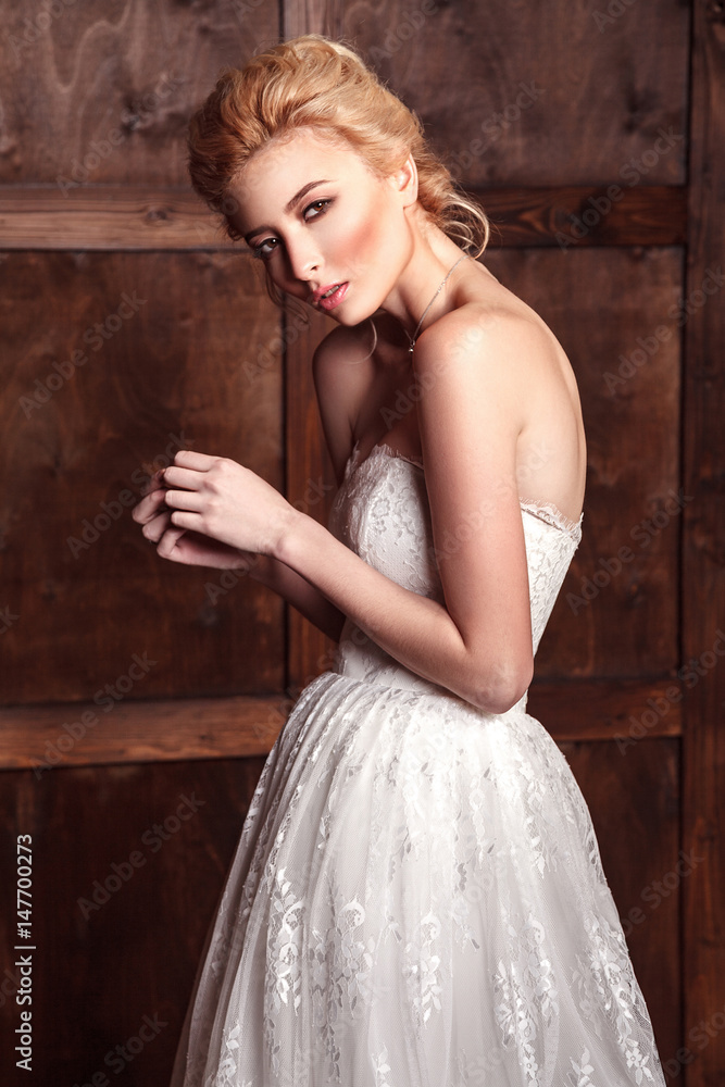 Fashion beauty bridal shooting. Beautiful fashion bride in wedding dress posing in front of woody background standing near brown leather sofa. studio shot in vogue style. 