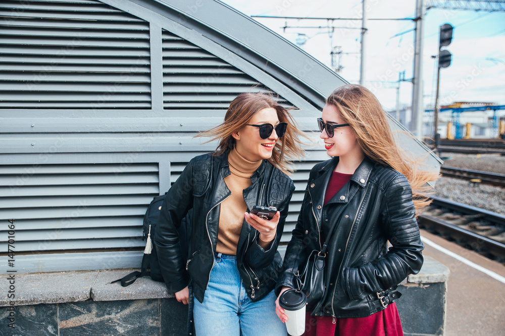 Fototapeta premium Two Women Talking in the City.Outdoor lifestyle portrait of two best friends hipster girls wearing stylish Leather Jacket and sunglasses with cofee, going crazy and having great time together