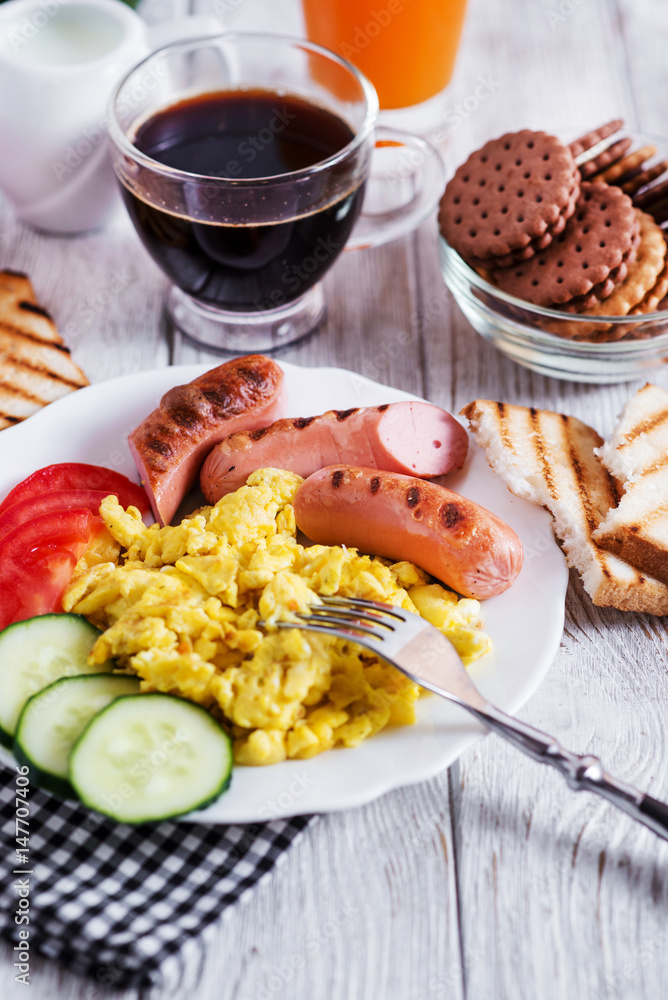 Hearty and traditional breakfast, scrambled eggs and milk with grilled pork, vegetables, tomato, cucumber, toast, juice and black coffee on a white wooden background Hearty and traditional breakfast, scrambled eggs and milk with grilled pork, vegetables, tomato, cucumber, toast, juice and black coffee on a white wooden background