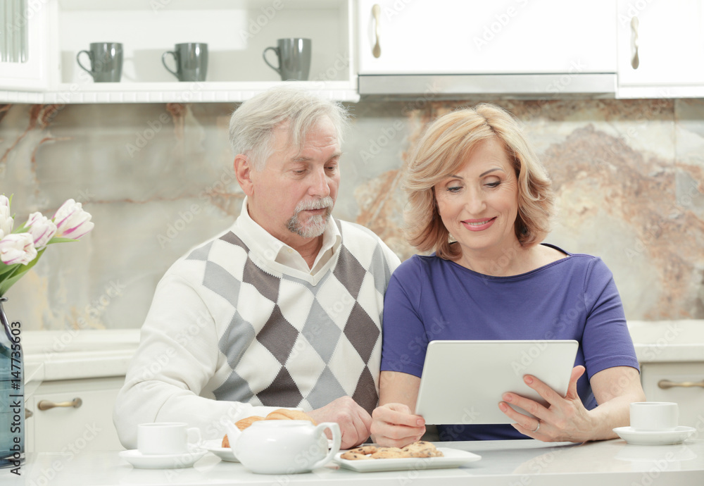 Cute elderly couple sitting in kitchen with tablet computer Cute elderly couple sitting in kitchen with tablet computer