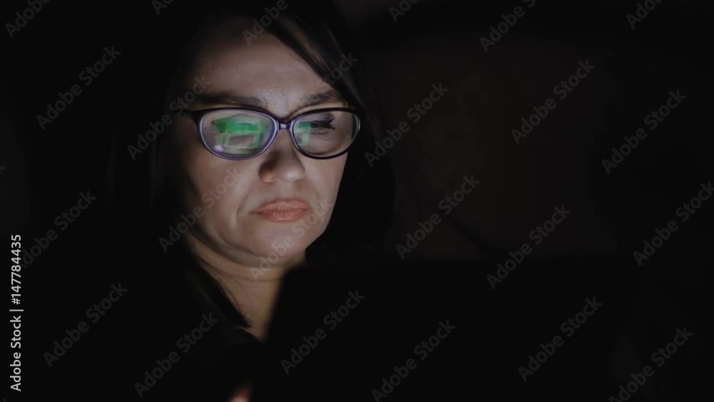 A woman is sitting on a sofa in a dark room and working on a tablet. A woman is sitting on a sofa in a dark room and working on a tablet.
