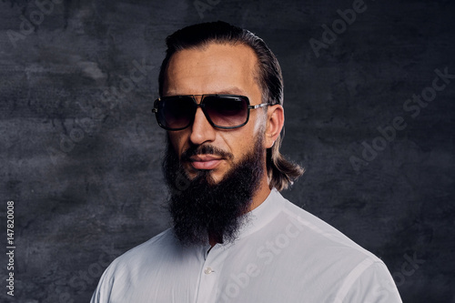 Bearded male with long hair dressed in a white shirt and sunglasses.