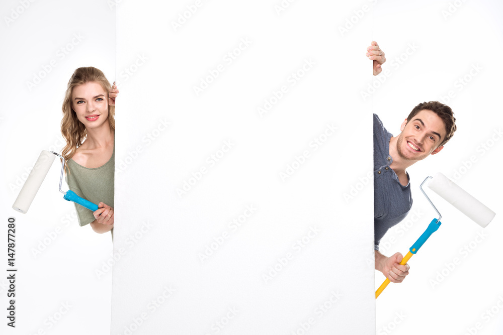 Smiling young couple with paint rollers and blank card looking at camera, renovation concept Smiling young couple with paint rollers and blank card looking at camera, renovation concept