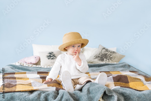 Pretty little girl in straw hat with blue eyes and a thoughtful expression sitting on her bed