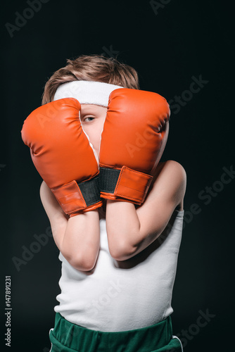 boy obscuring face with boxing gloves isolated on black, active kids concept