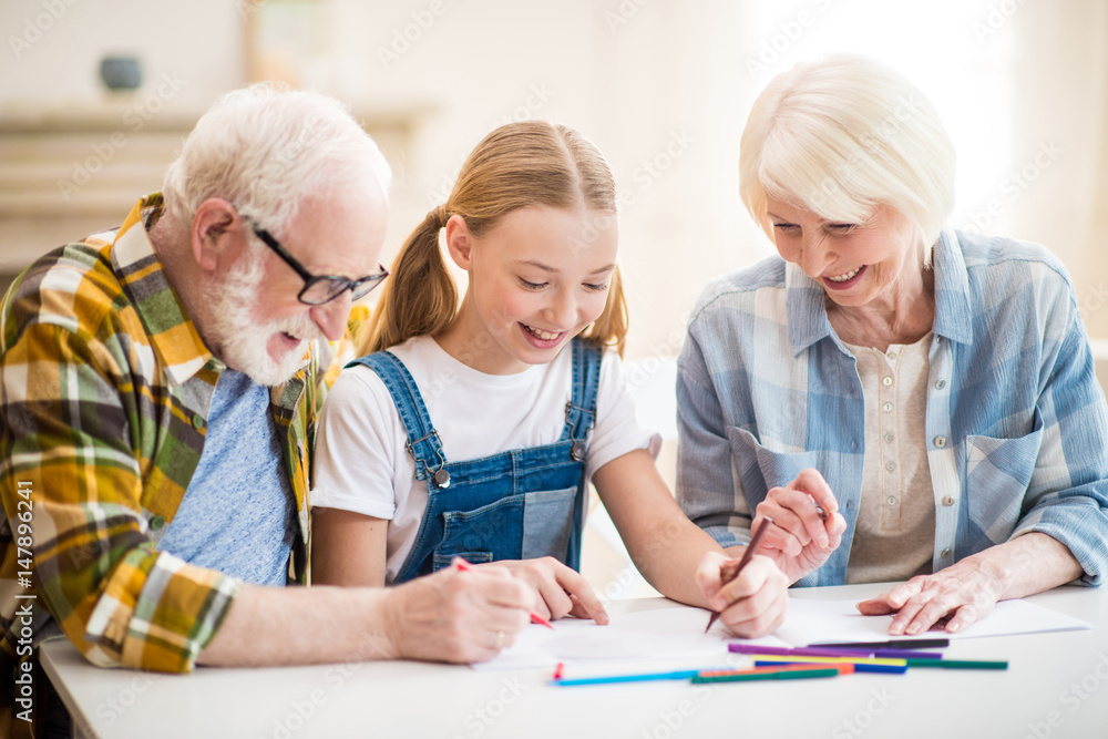Happy girl with grandfather and grandmother sitting at table and drawing together Happy girl with grandfather and grandmother sitting at table and drawing together