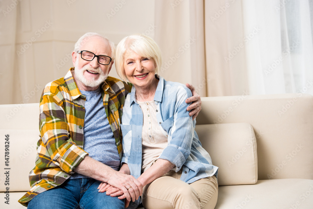 Happy senior couple sitting embracing on sofa and smiling at camera