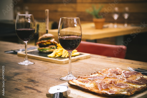 Traditional restaurant atmosphere with ham pizza and burger with chips in the background on rustic wooden table and glass of red wine