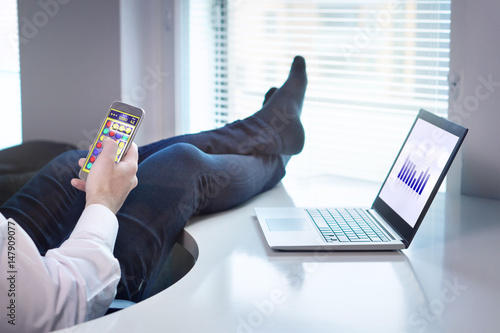 Lazy office worker playing mobile game with smartphone during work hours. Avoiding his job and being lazy with feet and socks on table. Useless and relaxing man doing nothing and forget his job. photo