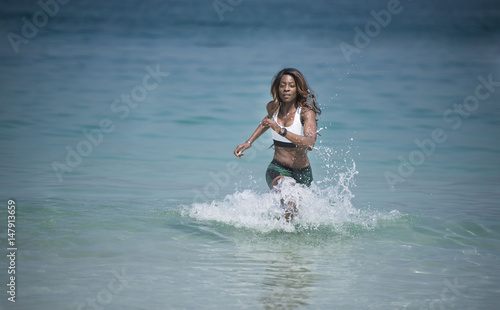 Beautiful African American Black athletic woman running out of the ocean wearing a sports bra and short tights splashing the water. 