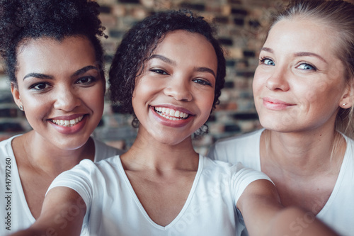 Beautiful and funny girls in white T-shirts makes selfie. Two afro-americans and one european.